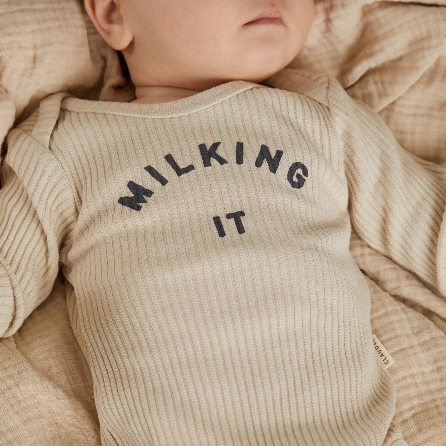Baby wearing a beige onesie with 'MILKING IT' text, lying on a soft surface.