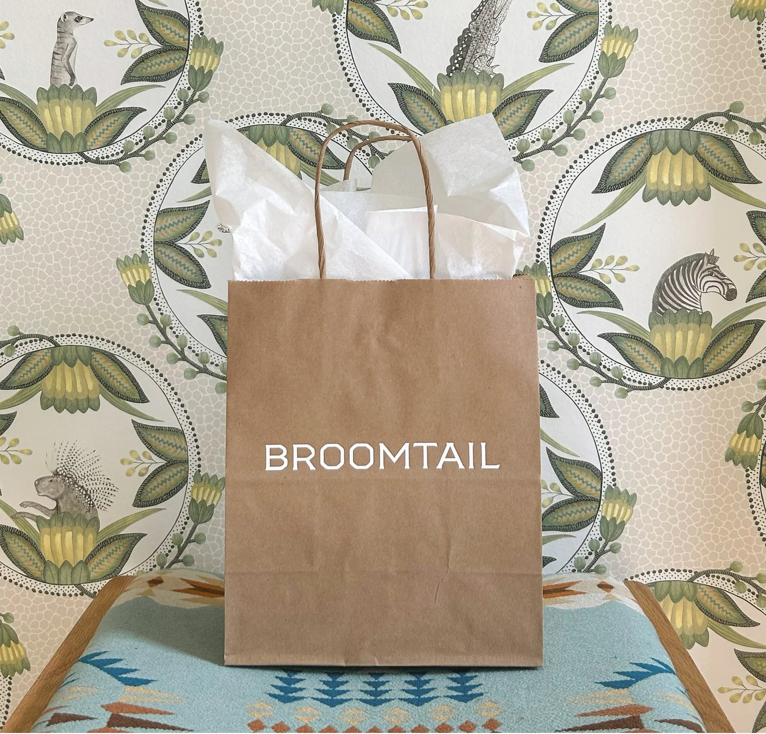 Brown paper bag with 'Broomtail' in front of the patterned wallpaper inside Broomtail boutique