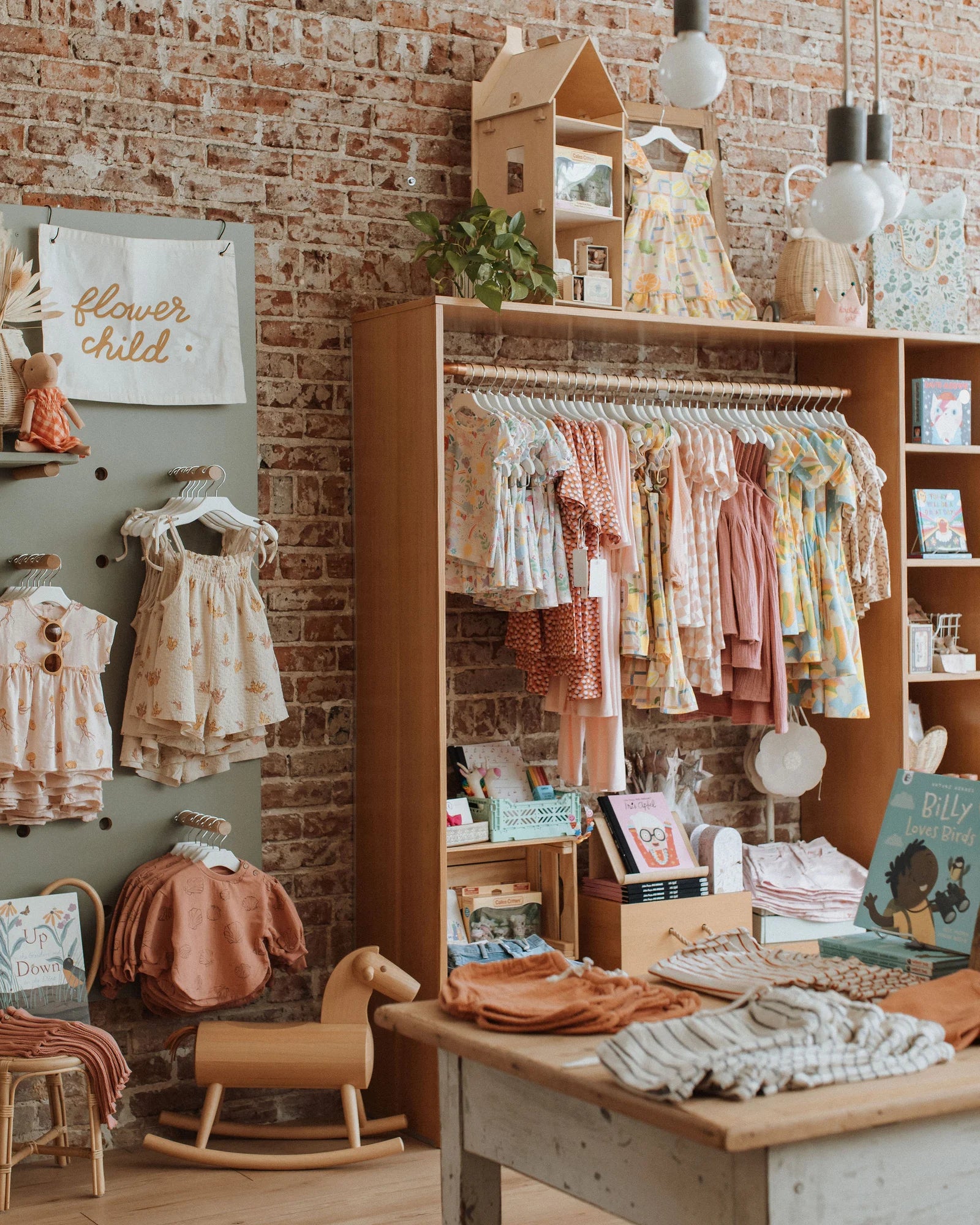 Baby and kids boutique clothing rack with handpicked dresses and books against a brick wall at Broomtail in Denver, Colorado.