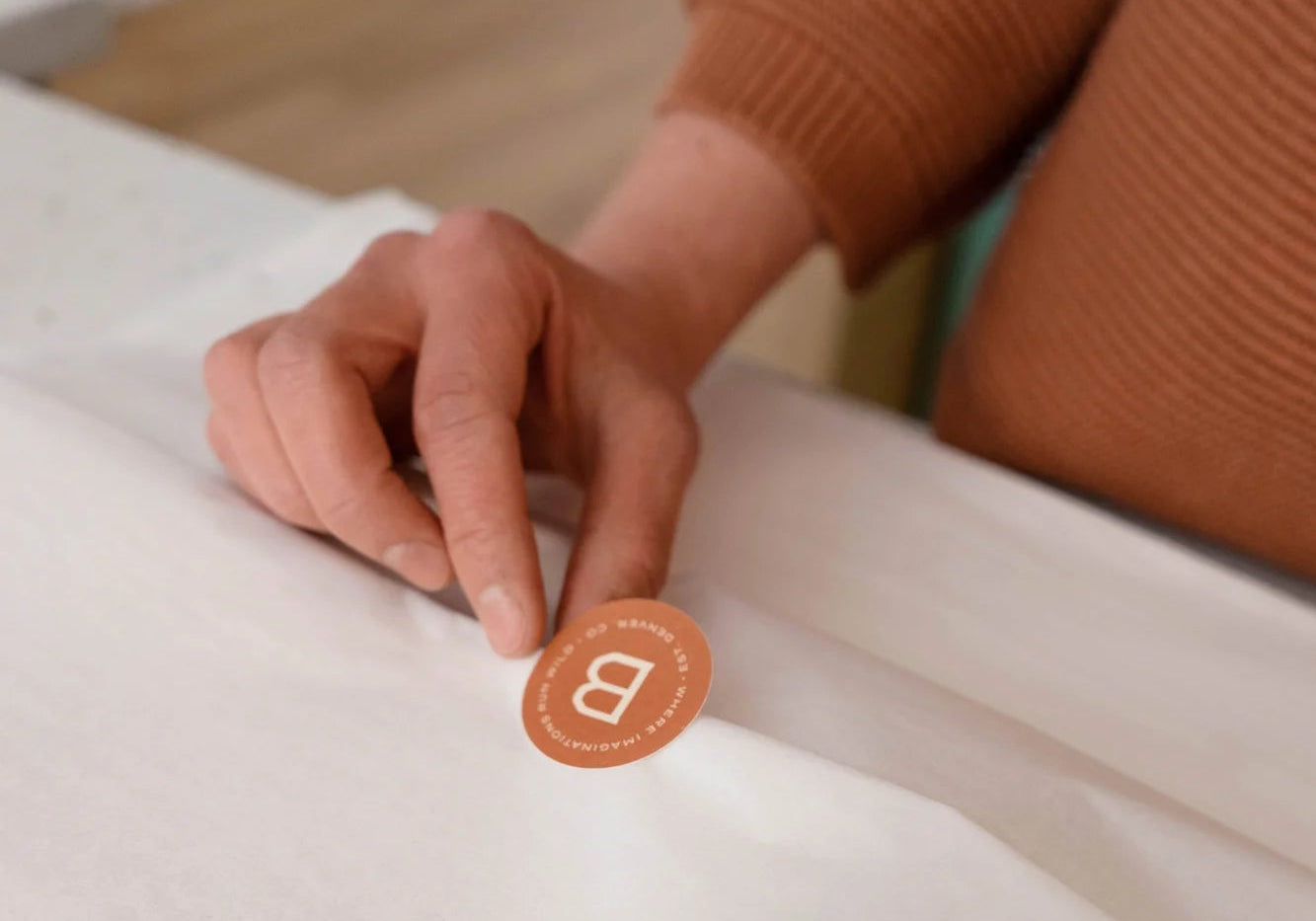 Hand holding a round orange sticker with a letter 'B' for "Broomtail" as part of the signature gift wrapping service offered in-store and online